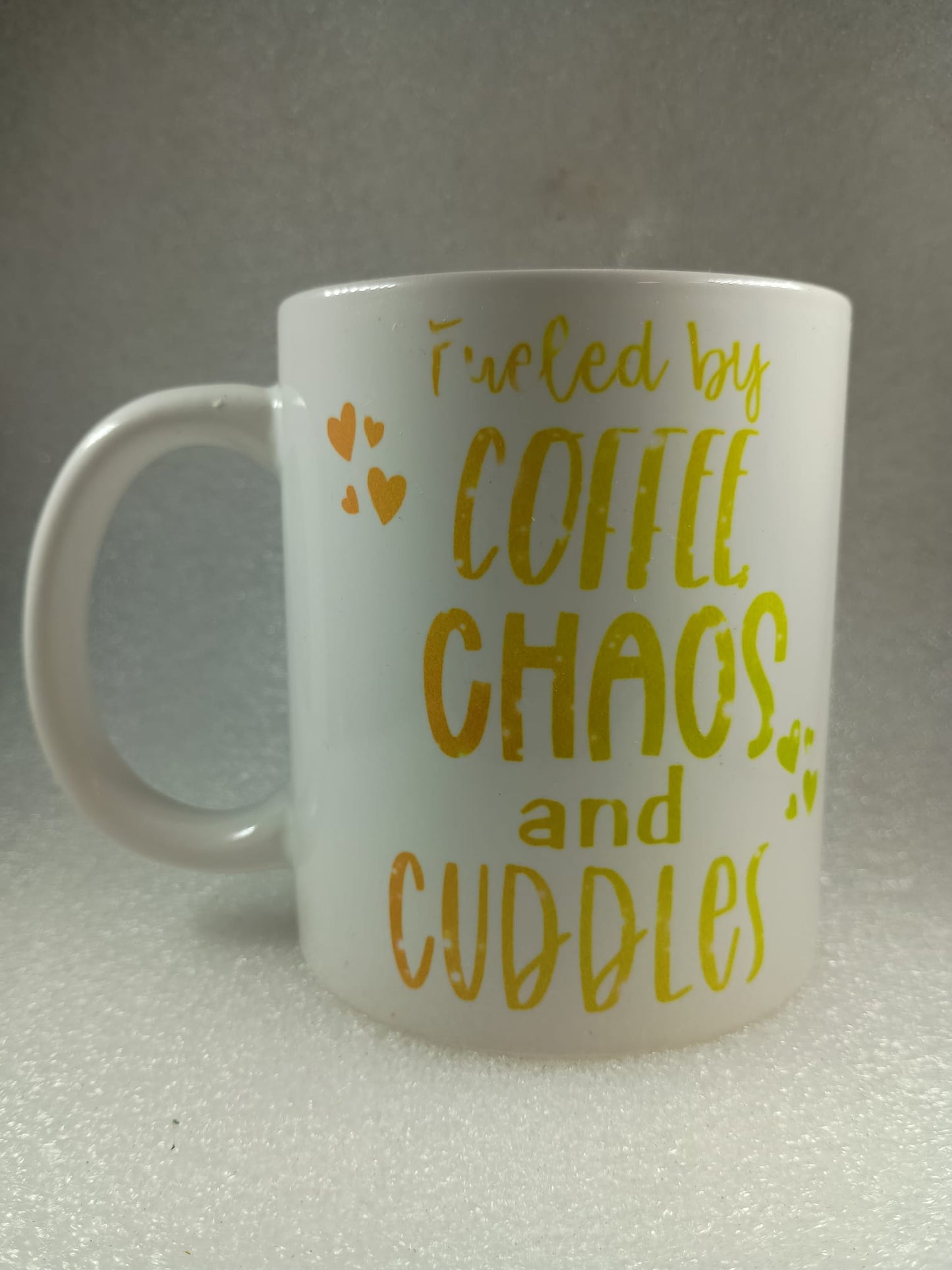 White mug with yellow text 'Fueled by coffee, chaos, and cuddles' on a gray background